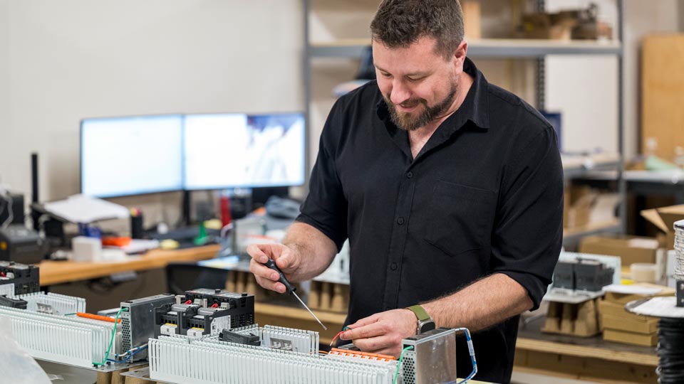 A man working with electronic equipment at a workstation, using a screwdriver and smiling while focused on his task.