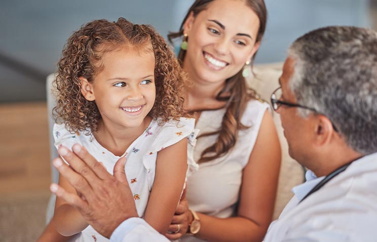 A doctor smiles and high-fives a young child while the child&rsquo;s mother looks on, conveying a warm, supportive health care visit.