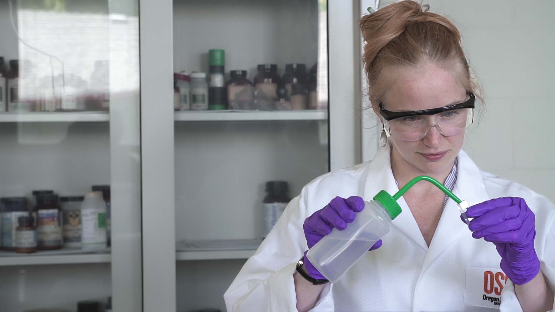 A woman in a lab coat, safety goggles and purple gloves uses a wash bottle in a laboratory with chemical containers visible in the background.