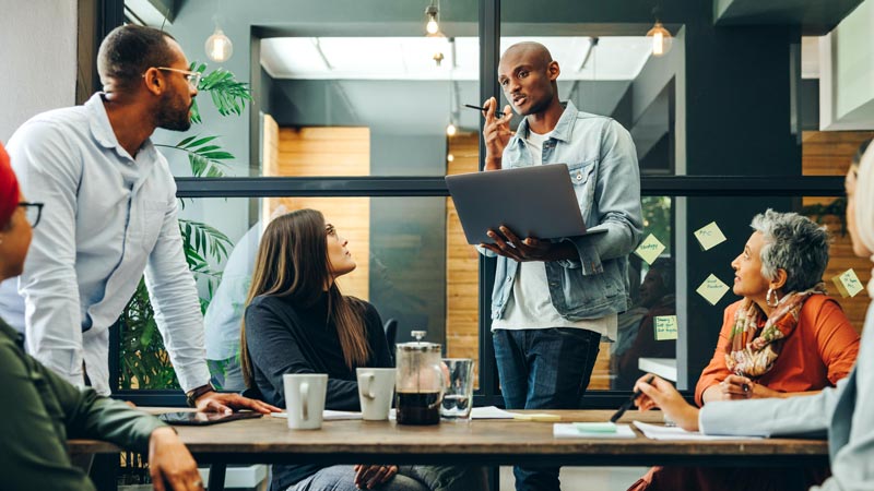 A man stands holding a laptop while speaking to five others gathered around a table in an office room.