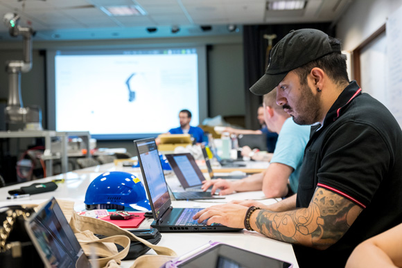 A group of people seated at a long table in a classroom or workshop setting, each working on laptops. In the background is a large screen that displays a presentation.