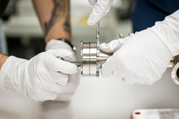 Two people wearing white gloves work together on a metallic cylindrical component. One person is holding a wrench, tightening or loosening a bolt, while the other stabilizes the pipe.