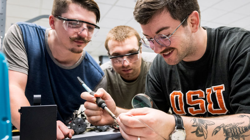 An Oregon State University student works with a soldering iron and magnifying glass in a training facility at Analog Devices Inc. Two other students lean over and look on.