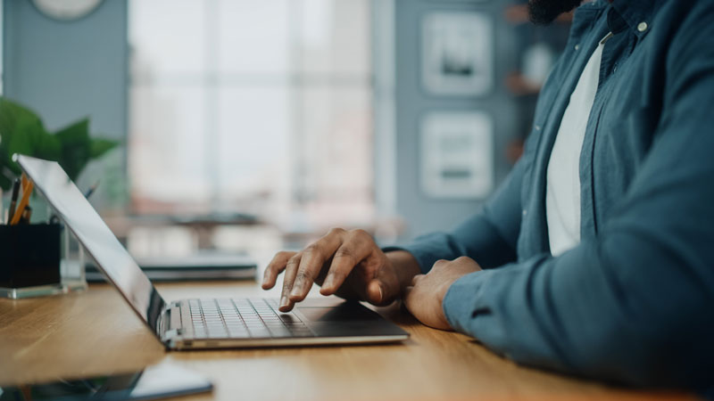 A person seated at a wooden desk using a laptop. The focus is on their hands and the device.