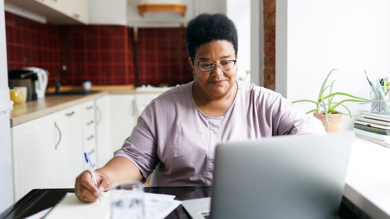 A woman sitting at a kitchen table looks at a laptop screen while writing in a notebook.