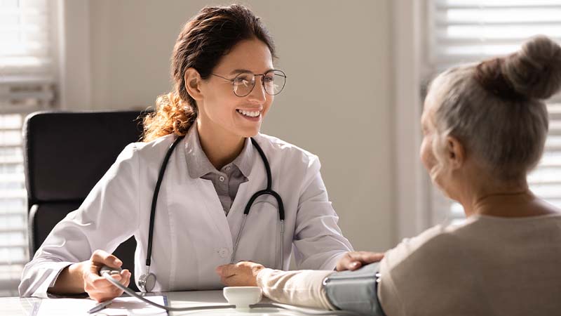 A person in a white coat wearing a stethoscope smiles across the desk at another person.