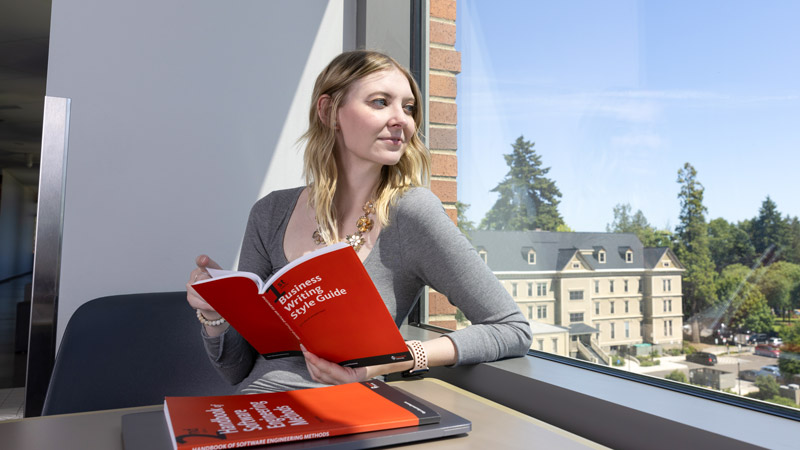 A woman reads a book titled "Business Writing Style Guide" by a window with a view of outside trees and a building.