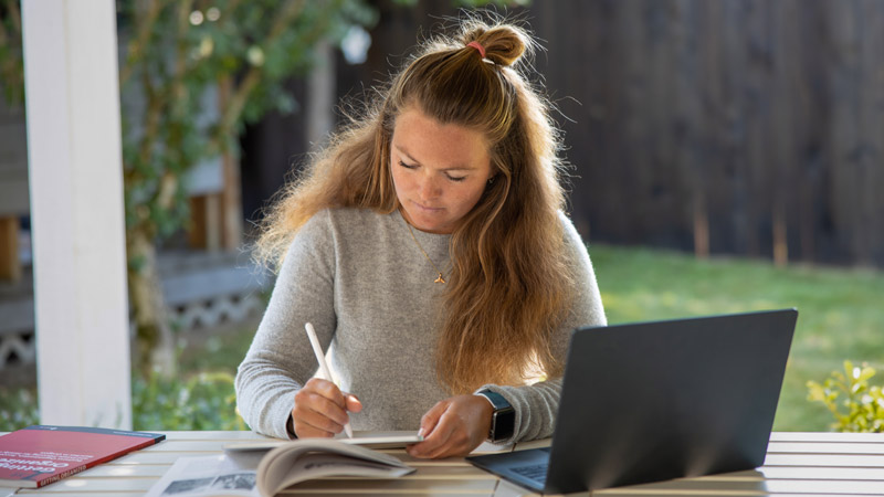A student sits at a desk in front of a window. A computer and papers sit on the desk. The student writes on the papers.
