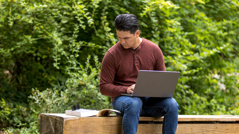 A student sits outside with a laptop and books. The student is looking down at the books.