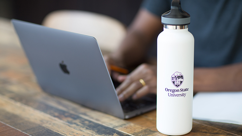 A person sitting at a table and typing on a laptop, with an Oregon State University water bottle in the foreground.